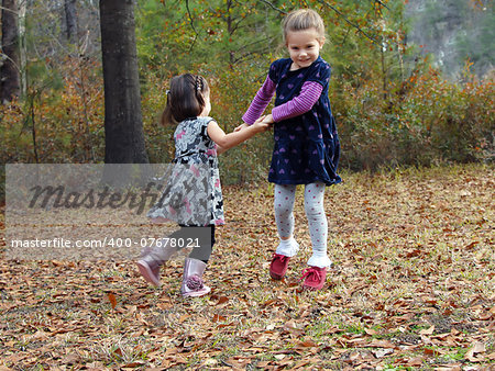 Two little girls hold hands and swing in circles.  They are playing outdoor games in the Fall leaves.