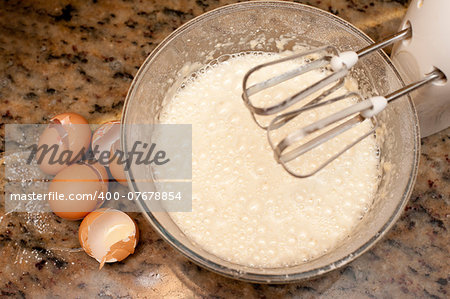 View from above of broken eggshells and a frothy batter mix in a metal mixing bowl on an electric mixer with whisk attachments for baking