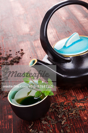 Traditional tea background. Tea in tea bowl with fresh mint herb, ceramic tea pot, dry tea crop on brown wooden background. Asian tea drinking ceremony.
