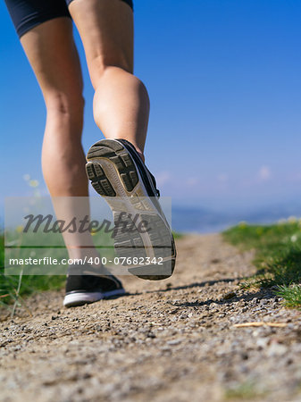 Photo of the legs and shoes of a young woman jogging on a gravel path down a country path.