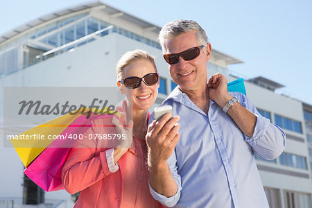 Happy senior couple looking at smartphone holding shopping bags on a sunny day
