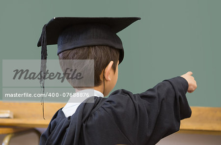 Cute pupil in graduation robe pointing in classroom at the elementary school