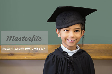 Cute pupil in graduation robe smiling at camera in classroom at the elementary school