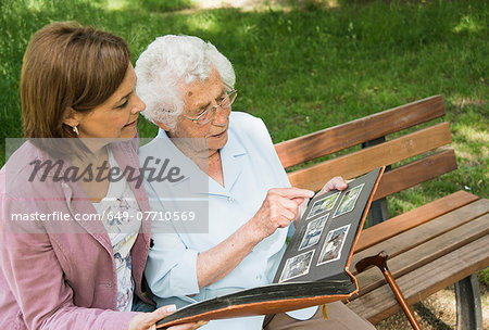 Senior woman sitting on park bench with granddaughter, looking at old photograph album