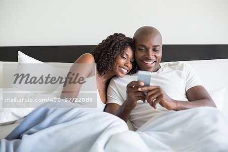 Happy couple cuddling in bed with smartphone at home in the bedroom