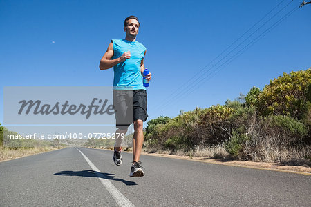 Athletic man jogging on open road holding bottle on a sunny day