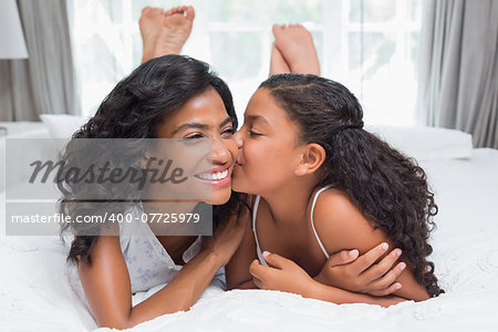 Pretty woman lying on bed with her daughter kissing cheek at home in bedroom