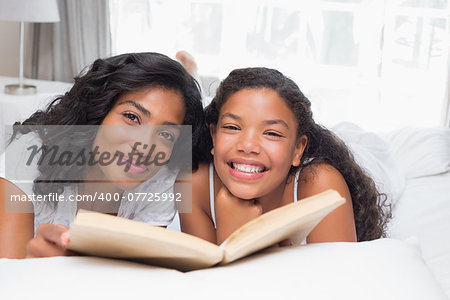 Mother and daughter reading book together on bed at home in bedroom