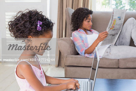 Cute daughter using laptop at desk with mother on couch at home in the living room