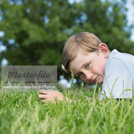 A young boy sitting on the grass using a hand held electronic games device.