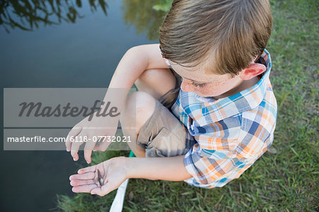 A young boy outdoors sitting on a riverbank with a small fish in the palm of his hand.