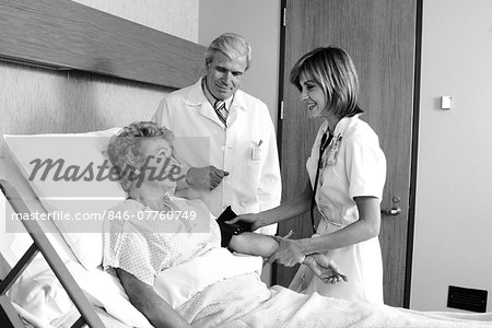 1980s ELDERLY WOMAN IN HOSPITAL BED HAVING BLOOD PRESSURE TAKEN BY FEMALE NURSE MALE DOCTOR STANDING BESIDE HER