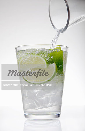 Clear water pouring from jug into drinking glass with lime and ice cubes Clear water pouring from jug into drinking glass with lime and ice cubes