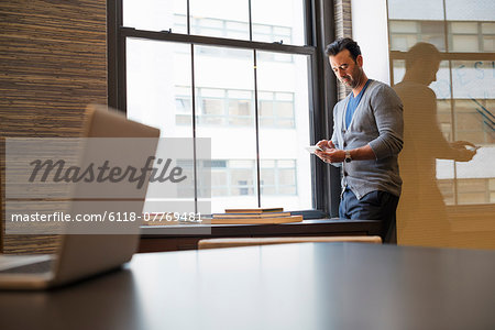 Office life. A man standing by a window in an office checking his smart phone.