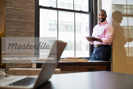 Office life. A man standing by a window in an office checking his smart phone.