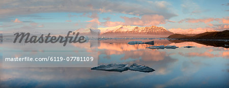 Panoramic view across the calm water of Jokulsarlon glacial lagoon towards snow-capped mountains and icebergs bathed in late afternoon light in winter, at the head of the Breidamerkurjokull Glacier on the edge of the Vatnajokull National Park, South Iceland, Iceland, Polar Regions