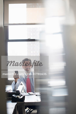 A man working at an office computer, holding a smart phone up to check for messages.