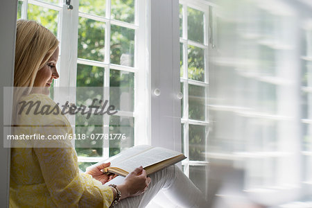 Woman sitting by a window, reading a book.