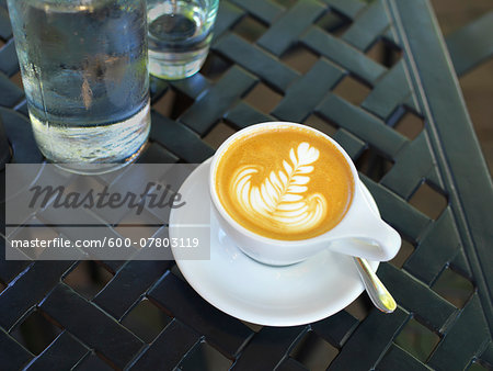 Close-up of cappuccino with latte art in white cup and saucer with water bottle and glass on patio table, Canada Close-up of cappuccino with latte art in white cup and saucer with water bottle and glass on patio table, Canada