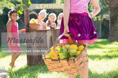 Cropped shot of girl carrying basket of lemons in front of  lemonade stand