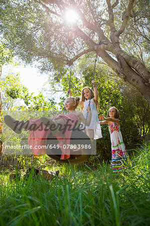 Low angle view of three girls playing on tree tire swing in garden