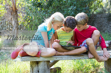 Three children sitting on garden seat looking down at digital tablet