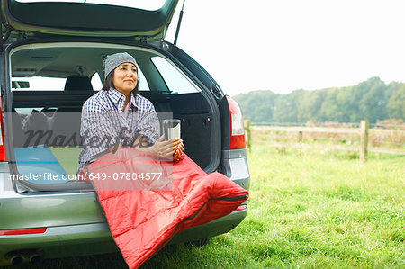 Woman sitting at rear of car with legs tucked in sleeping bag