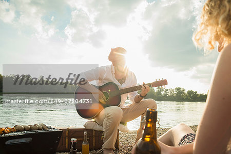 Young man sitting by lake playing guitar