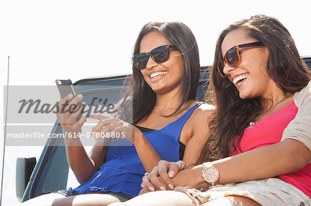 Two young women sitting on jeep hood looking at smartphone