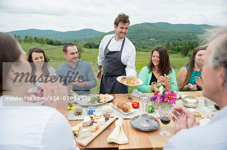 Mid adult man in apron, serving plate of food to family members at table, outdoors