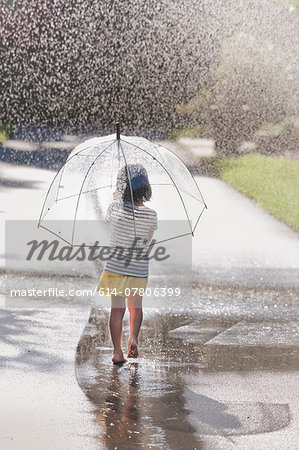Rear view of barefoot girl carrying umbrella walking through street puddle