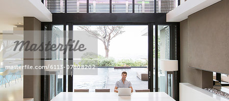 Young man using laptop in modern dining room with patio doors and swimming pool in background