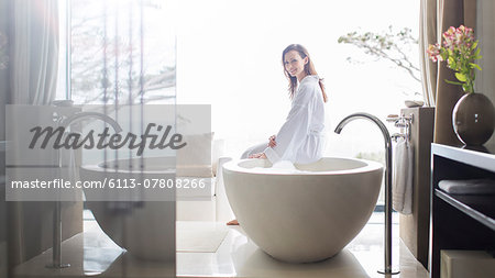 Portrait of smiling woman wearing white bathrobe, sitting on edge of bathtub in bathroom