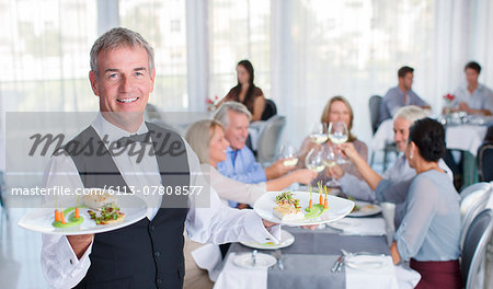Portrait of waiter holding plate with fancy meals, people at restaurant tables in background