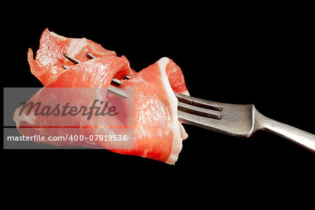 Delicious culinary prosciutto ham on silver fork isolated on black background. Culinary appetizer eating.