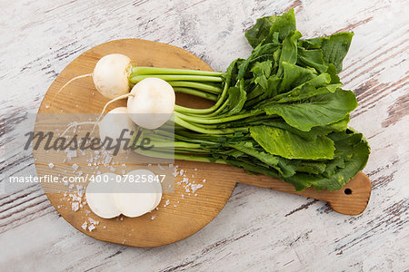 White round daikon radish with leaves on wooden cutting board on white wooden textured background. Culinary healthy country style vegetable still life.