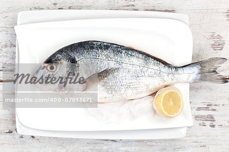 Fresh fish on white plate with lemon on white textured wooden background, top view. Culinary seafood concept.