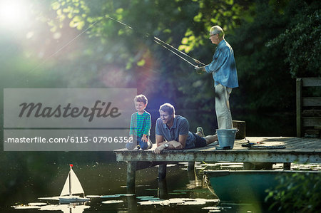 Boy fishing and playing with toy sailboat with father and grandfather at lake