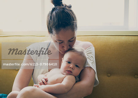 Portrait of mother holding and kissing baby, sitting on sofa by window