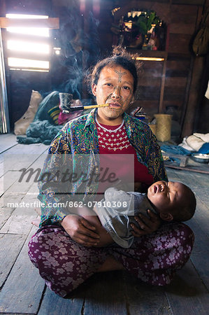 Myanmar, Chin State, Kempetlet. Chin woman with  traditional facial tattoo, holding her baby boy and smoking pipe. (MR) (PR)