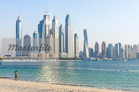 United Arab Emirates, Dubai. Dubai marina cityscape at sunset, with woman walking on the beach (MR)