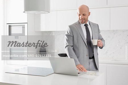 Mid adult businessman having coffee while using laptop in kitchen