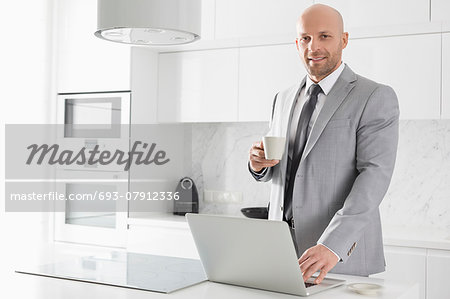 Portrait of confident mid adult businessman having coffee while using laptop in kitchen