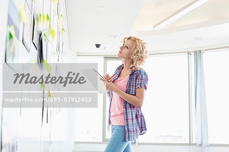 Creative businesswoman looking at papers stuck on wall while writing notes in office