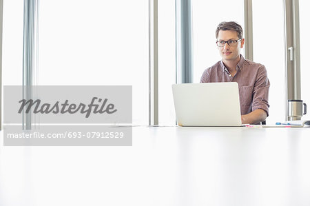 Thoughtful businessman looking away while using laptop at desk in creative office