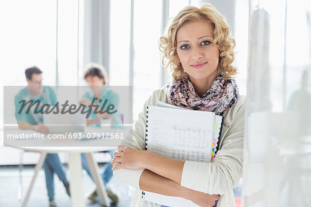 Portrait of creative businesswoman holding files with colleagues working in background at office