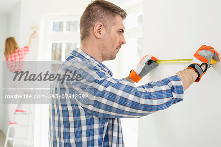 Man measuring wall with woman painting in background