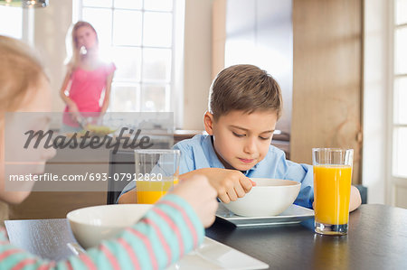 Siblings having breakfast at table with mother preparing food in background