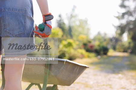 Midsection rear view of female gardener pushing wheelbarrow at plant nursery