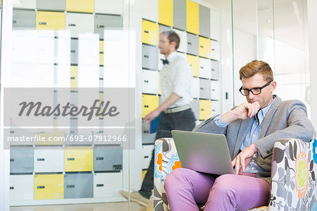 Businessman using laptop on armchair with colleague walking in background at creative office